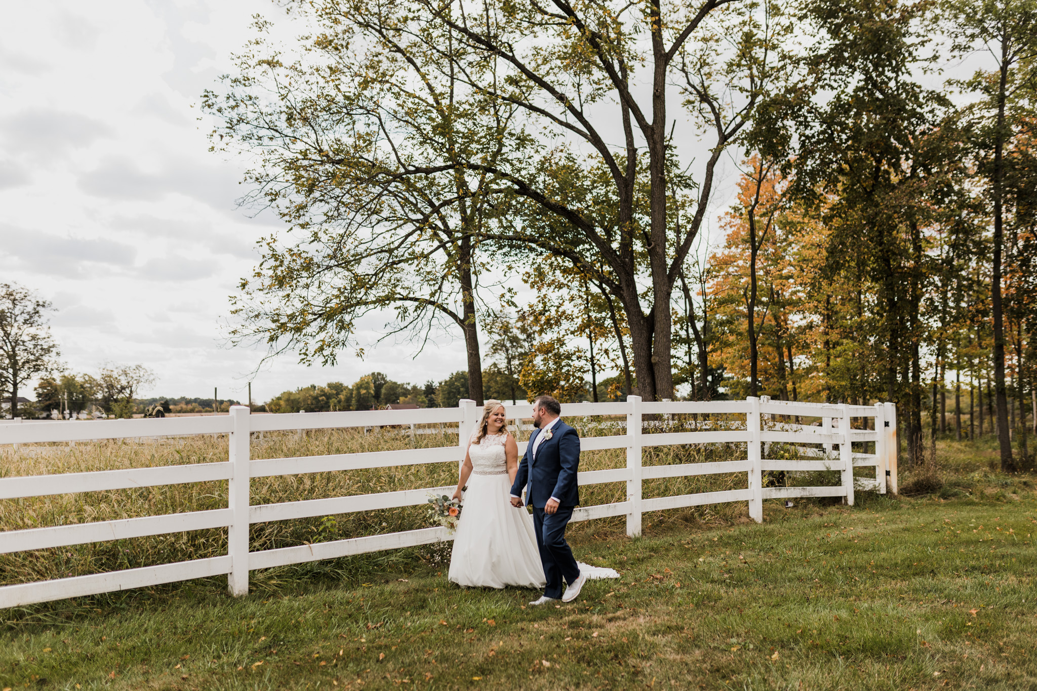 A bride and groom walk along the fence of a horse farm pasture during their Indiana wedding.