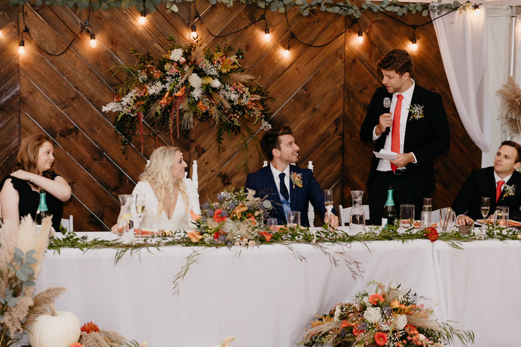 Groom and bride sitting at table, looking at best man give a speech in a white tent wedding