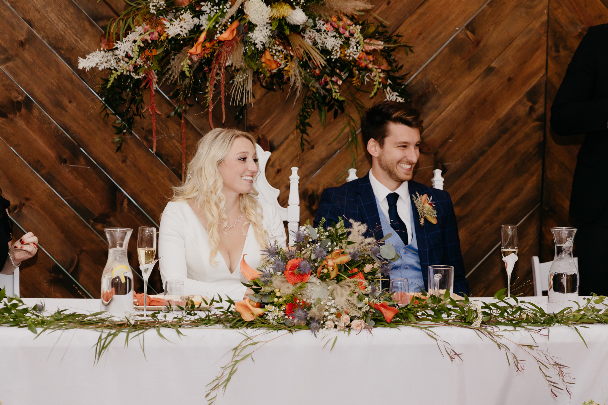 Groom and bride sitting at table, looking at best man give a speech in a white tent wedding
