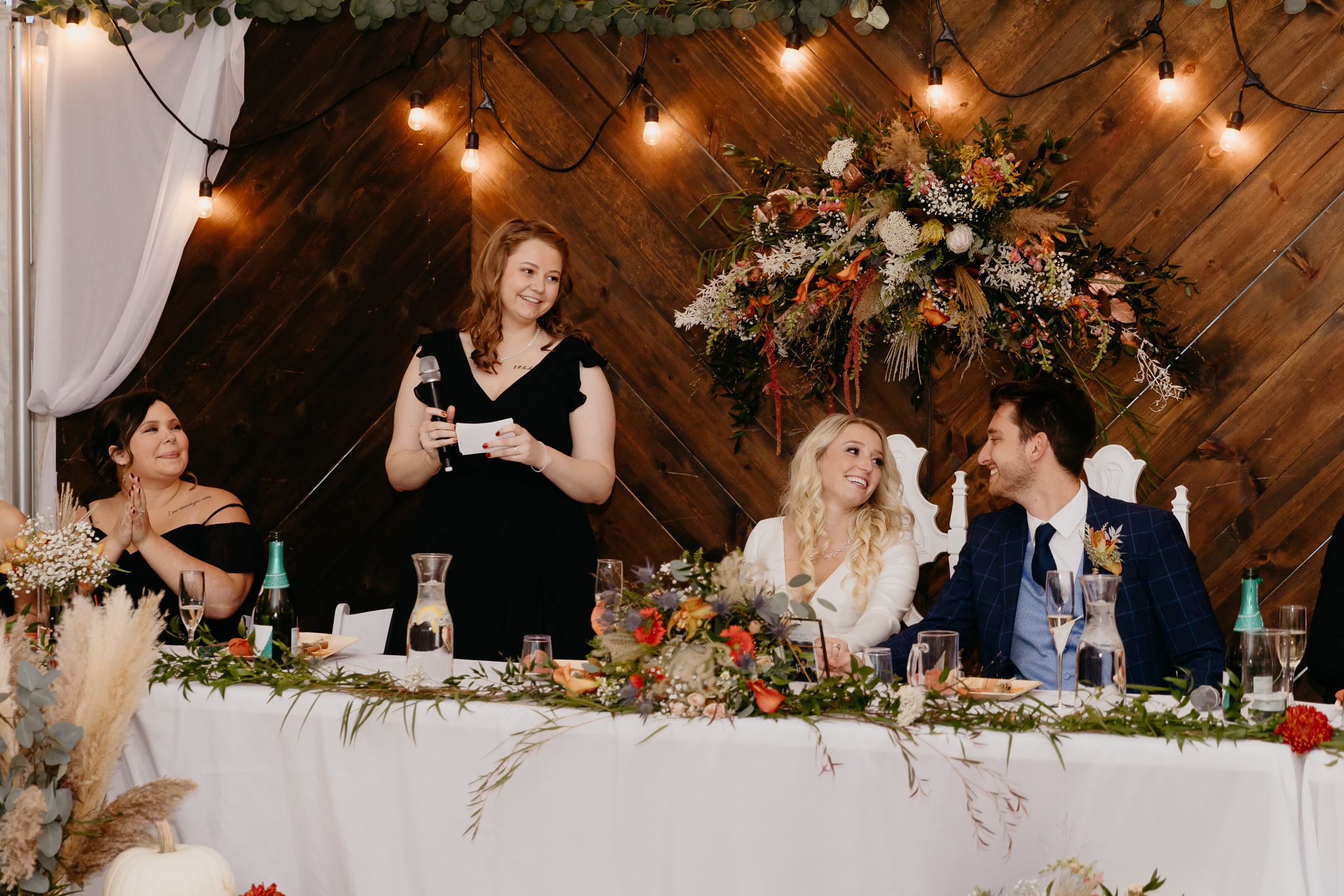 Groom and bride sitting at table, toast the maid of honor after giving a speech in a white tent wedding