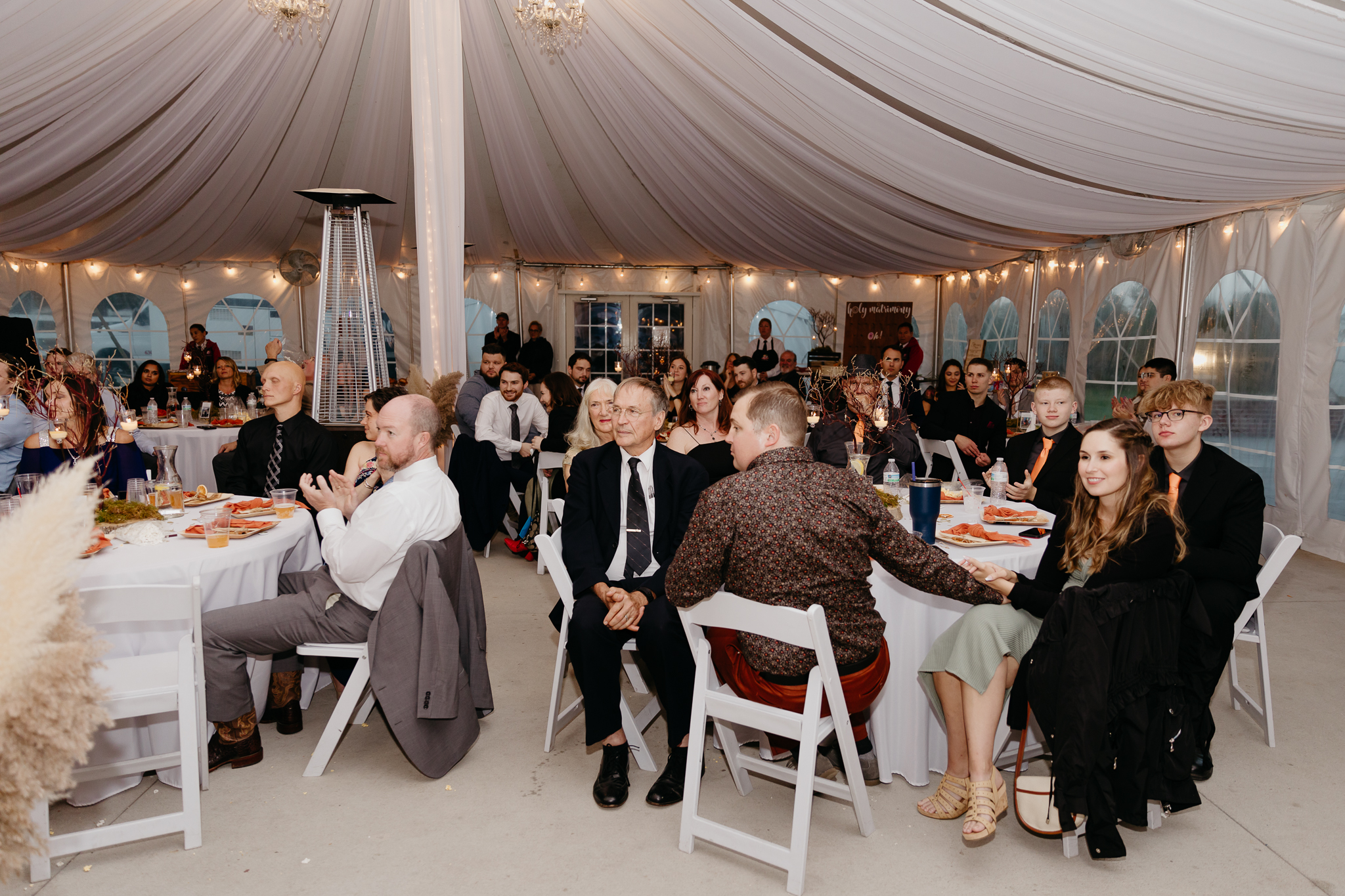 Wedding guests sit at round tables watching speeches in a white tent wedding