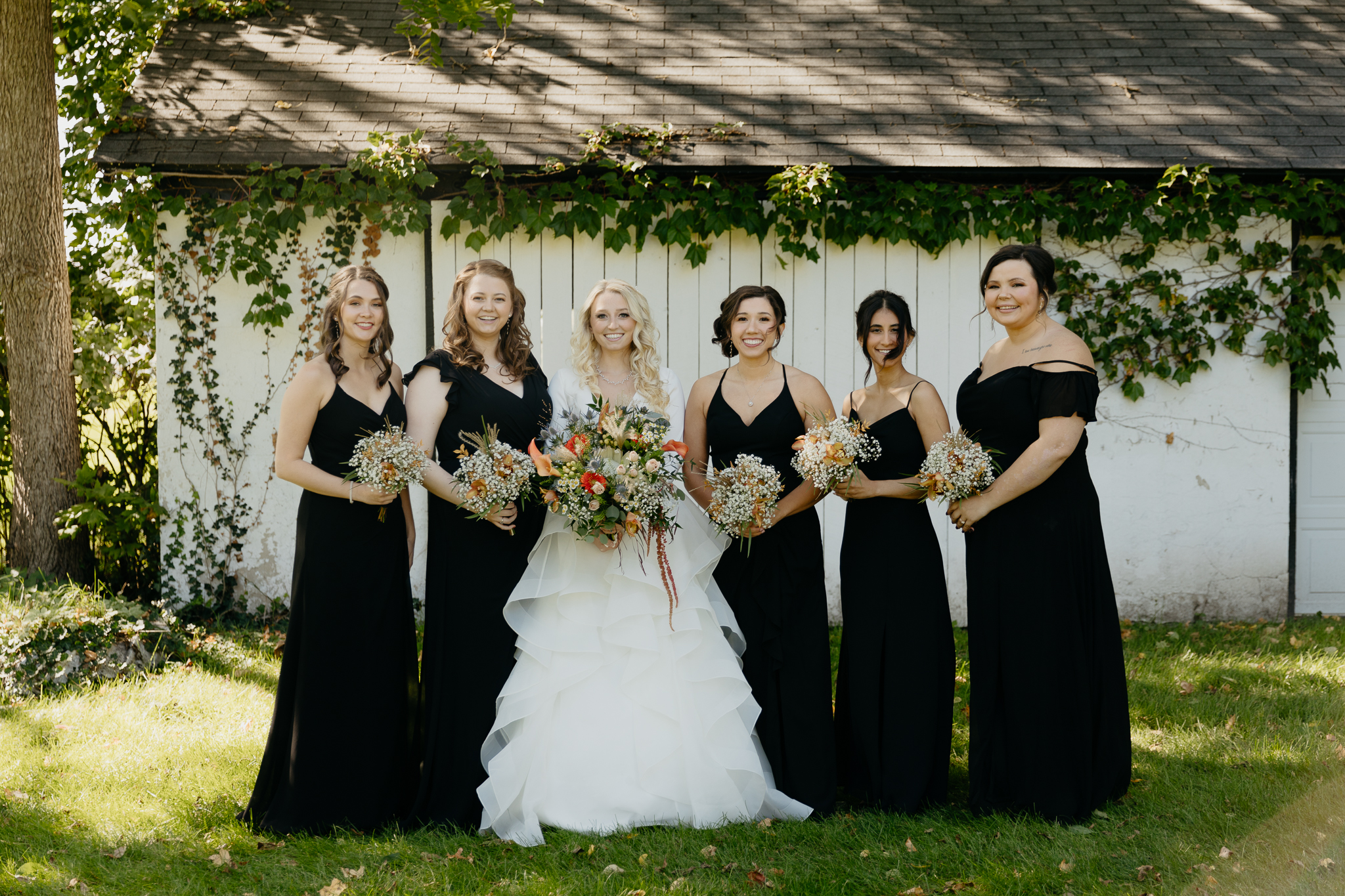 Bridesmaids and bride smile together with bouquets, standing in front of an ivy covered white barn.
