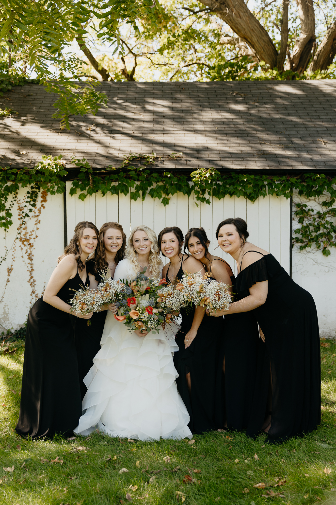 Bridesmaids and bride smile and laugh together with bouquets, standing in front of an ivy covered white barn at Northfork Farm