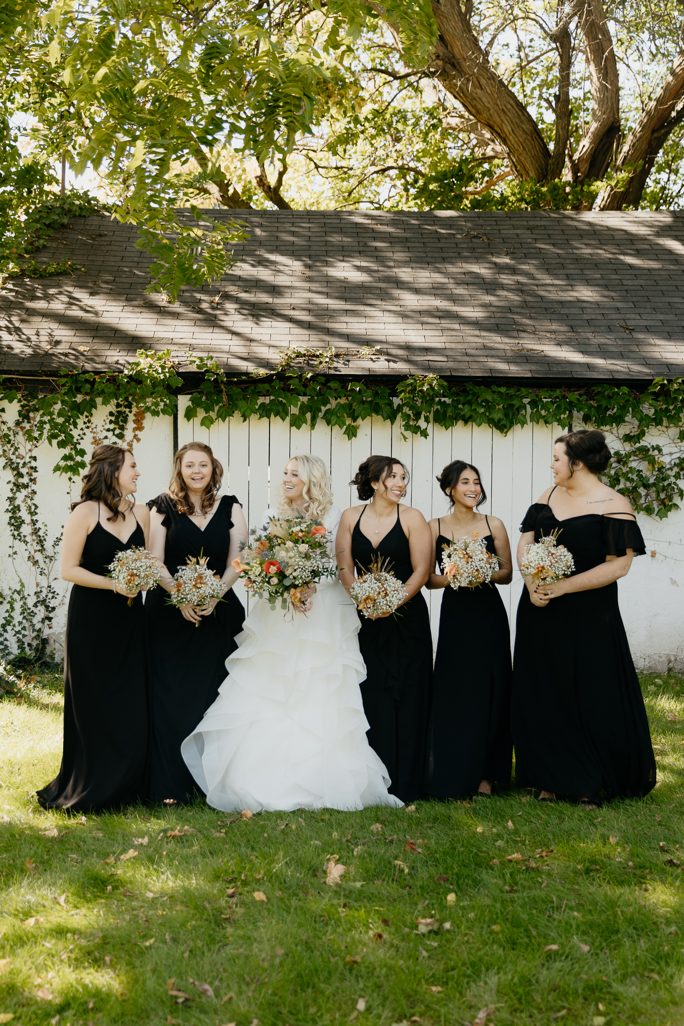 Bridesmaids and bride smile and laugh together with bouquets, standing in front of an ivy covered white barn at Northfork Farm