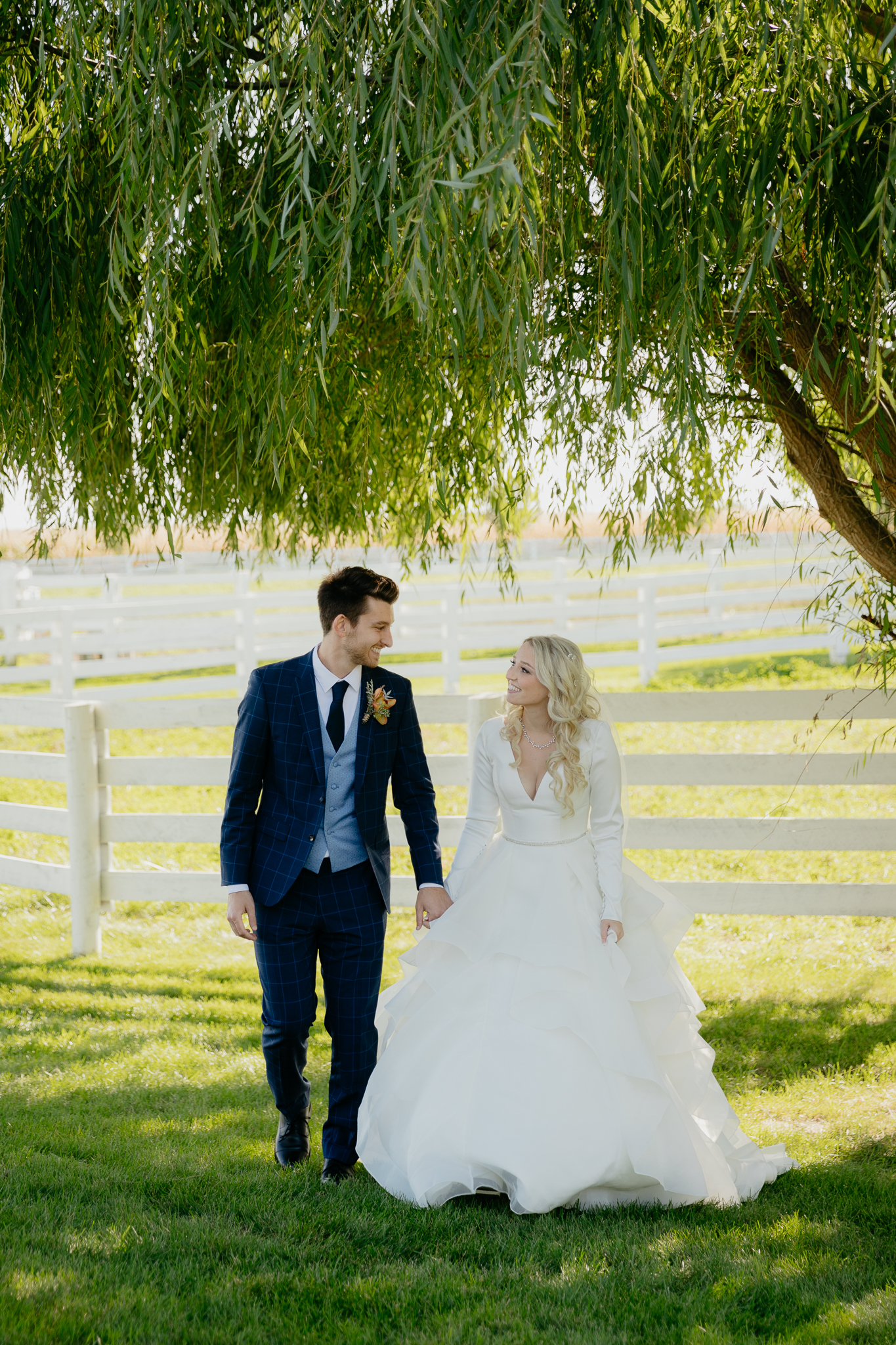 groom and bride walk hand in hand underneath a willow tree