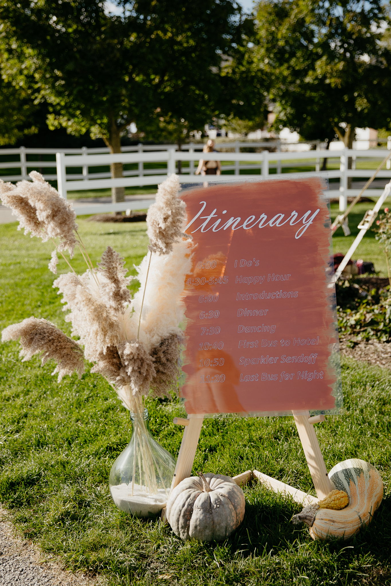 An orange wedding itinerary sign on the grass next to pampas grass