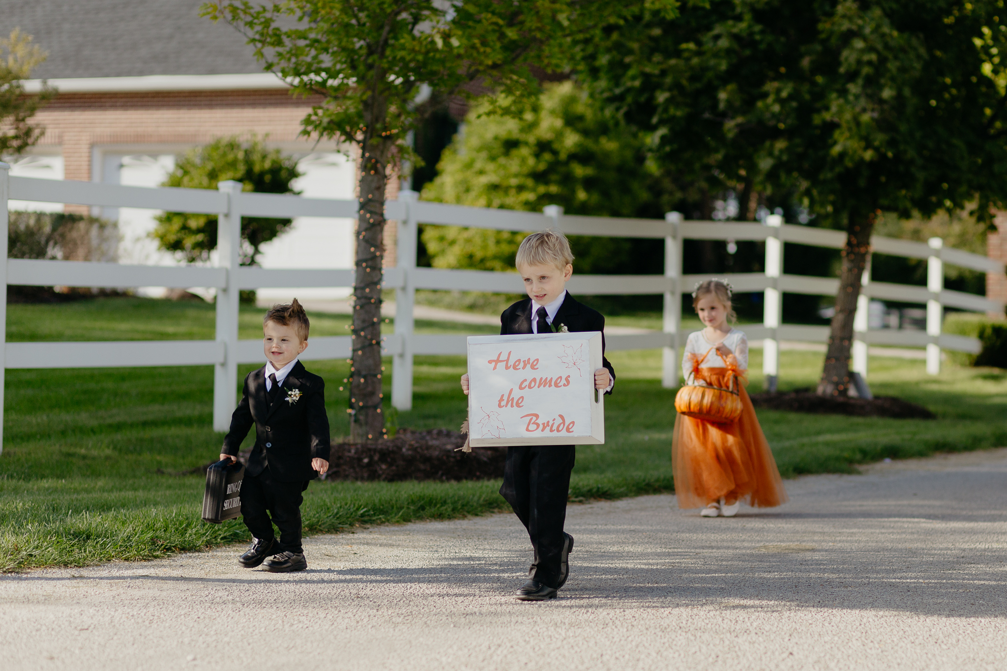 Flower girl and ring boys walk down tree lined driveway with a here comes the bride sign