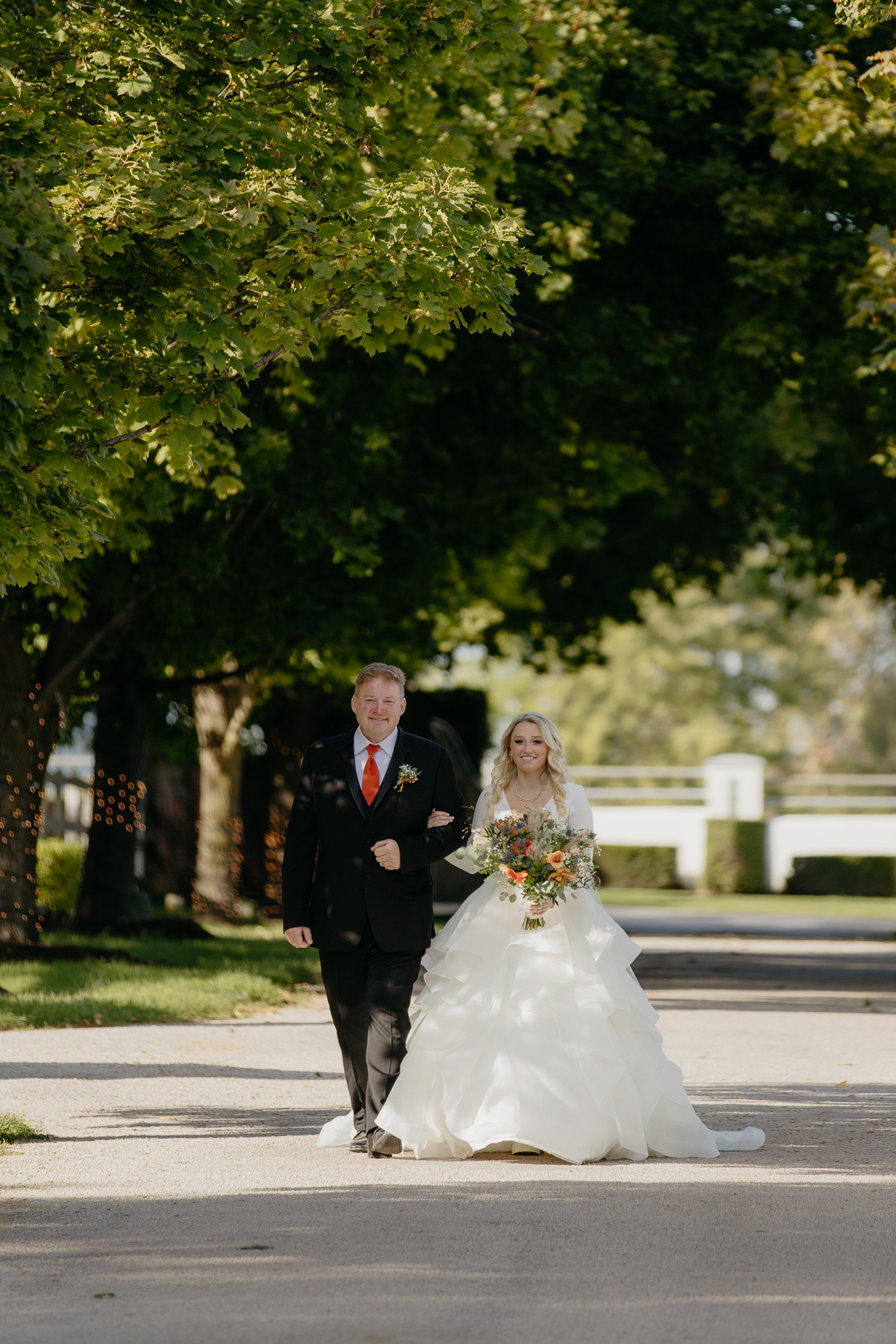 Bride in wedding dress walking down tree lined driveway arm in arm with her father