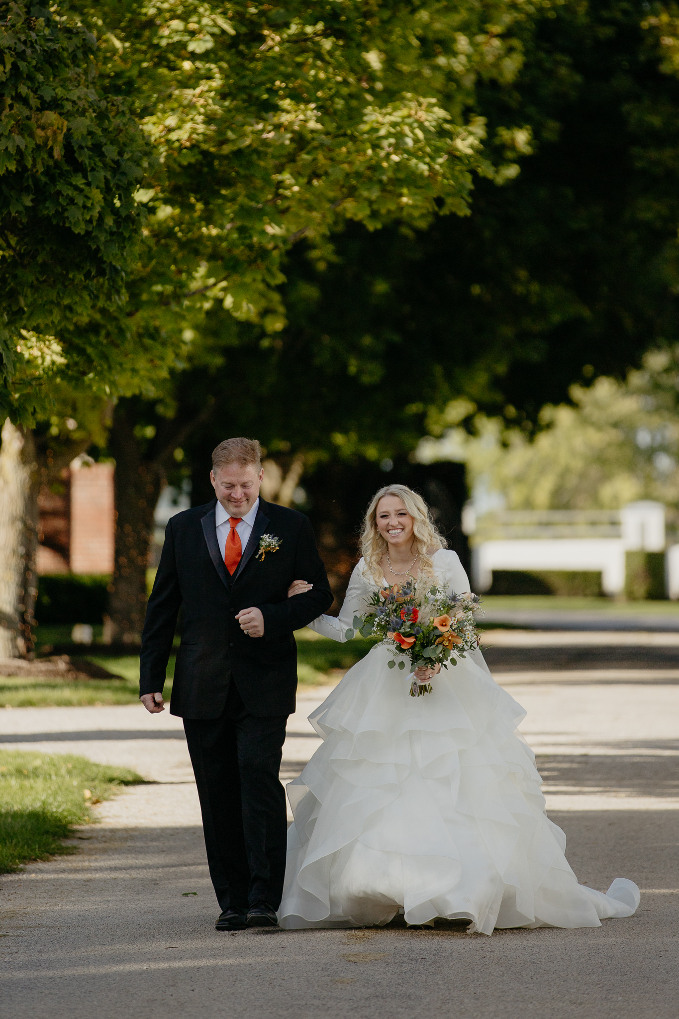 Bride in wedding dress walking down tree lined driveway arm in arm with her father