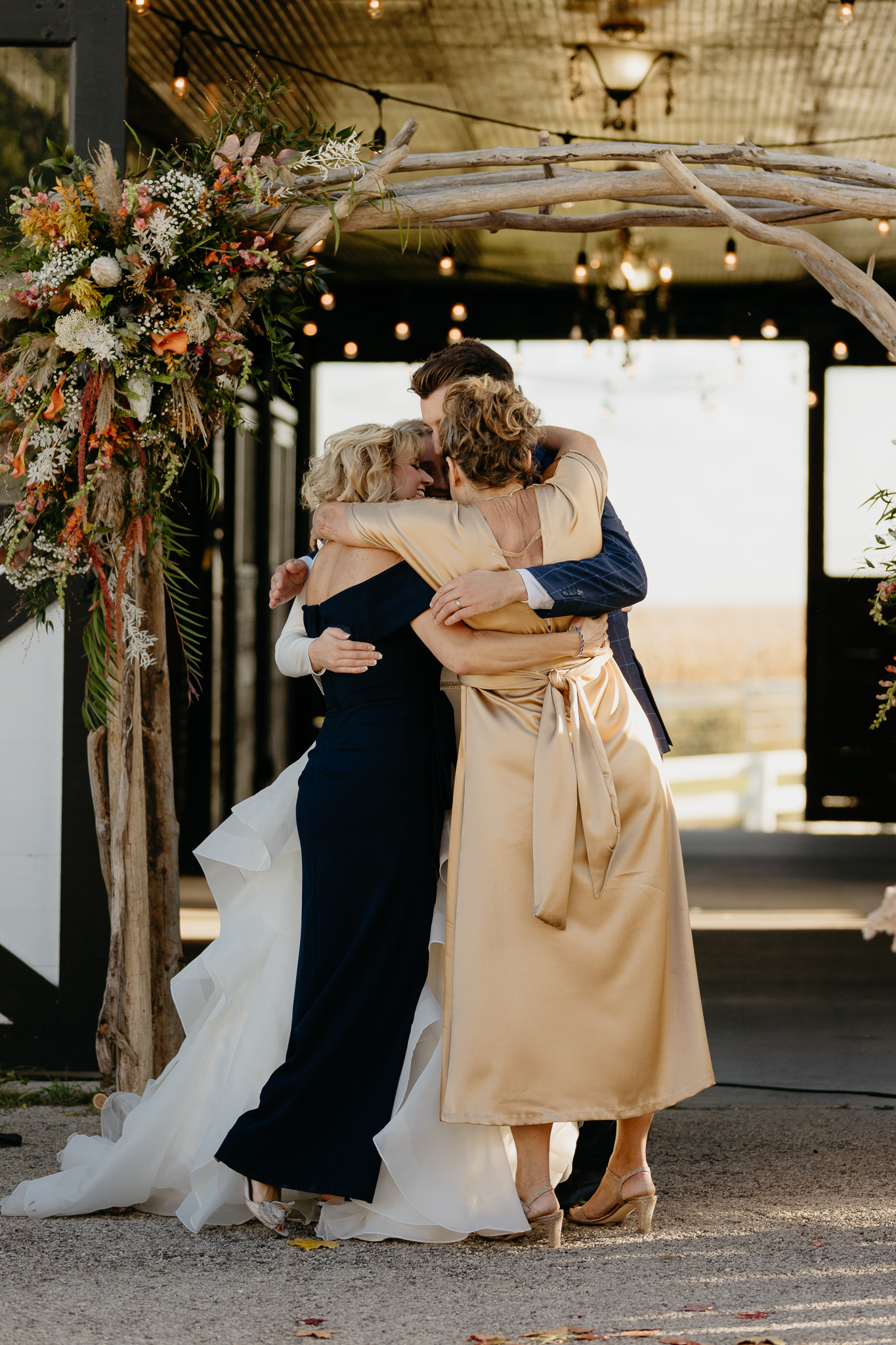 Bride, groom, and moms all hug at the altar of an outdoor wedding ceremony, in front of a white barn