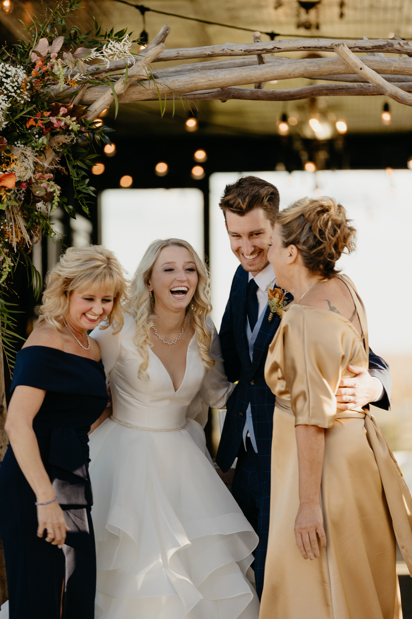 Bride, groom, and moms all hug and laugh at the altar of an outdoor wedding ceremony, in front of a white barn