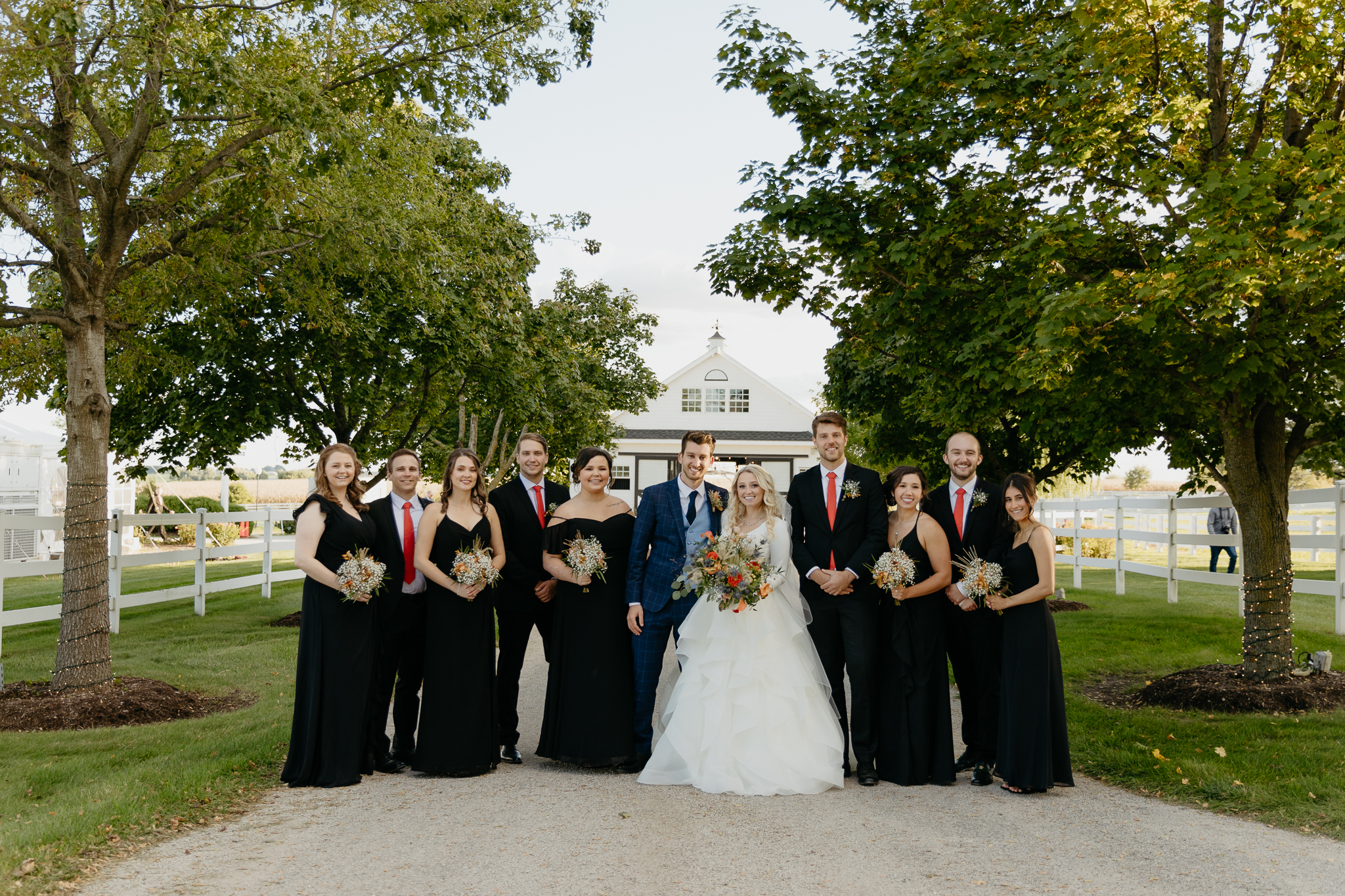 Wedding party of bridesmaids and groomsmen stand in a line and smile in front of a white horse stable and trees