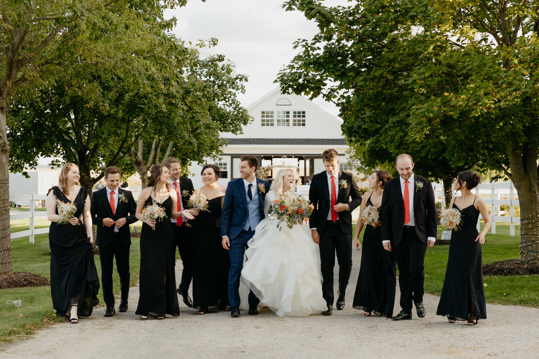 Wedding party of bridesmaids and groomsmen stand in a line and smile in front of a white horse stable and trees