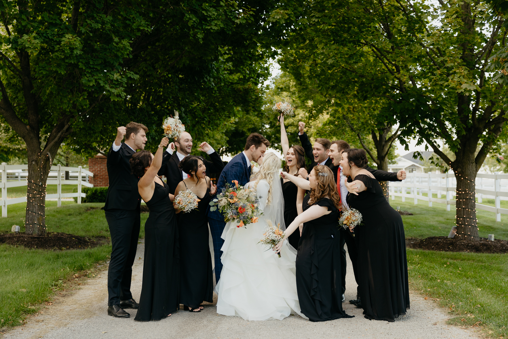 Wedding party of bridesmaids and groomsmen cheer as groom and bride kiss in a tree lined driveway at Northfork Farm