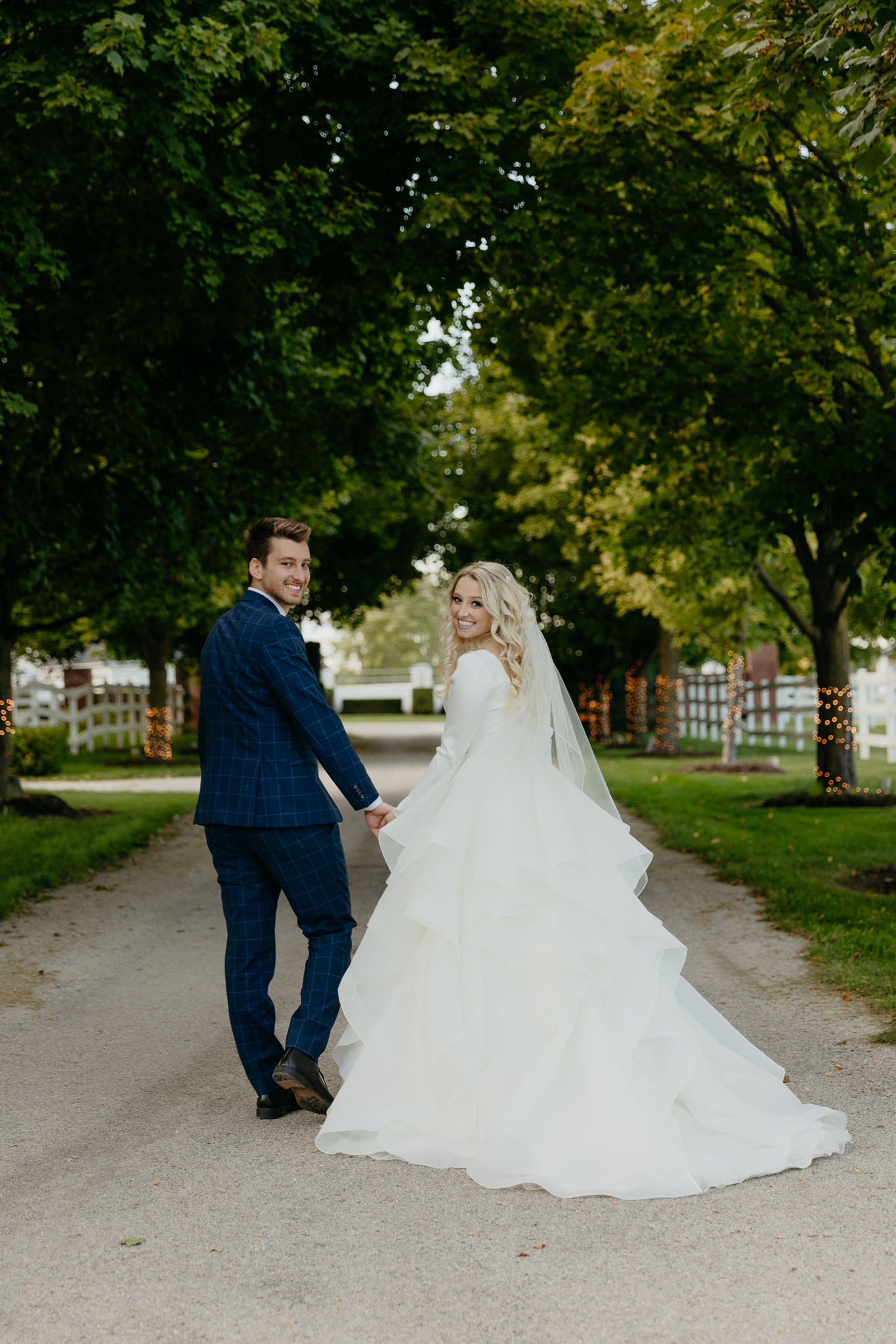 Bride and groom walk down tree lined driveway hand in hand while looking back
