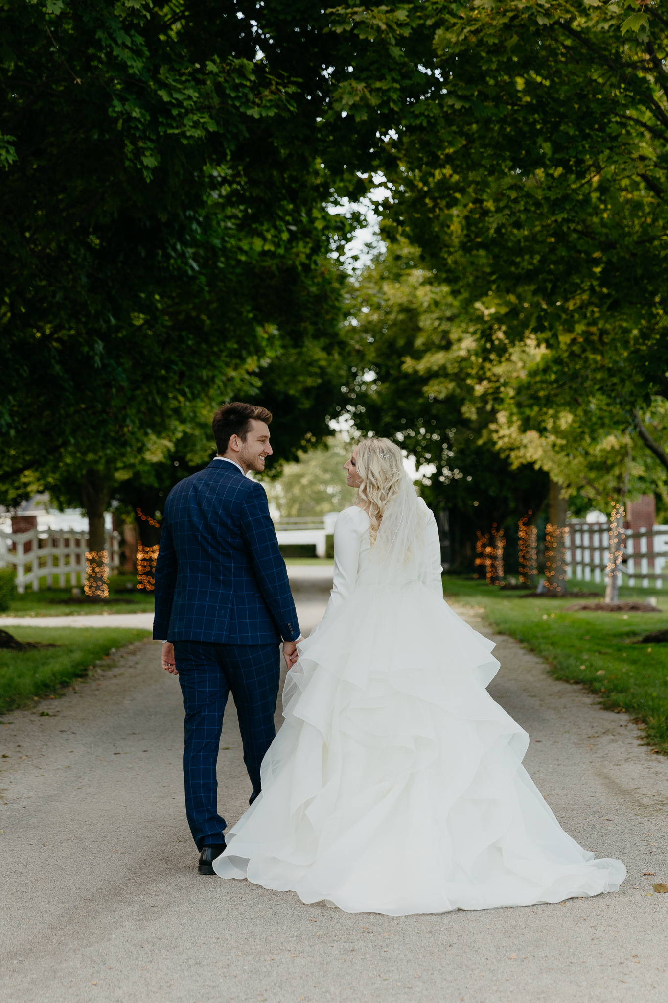 Bride and groom walk down tree lined driveway hand in hand while looking back