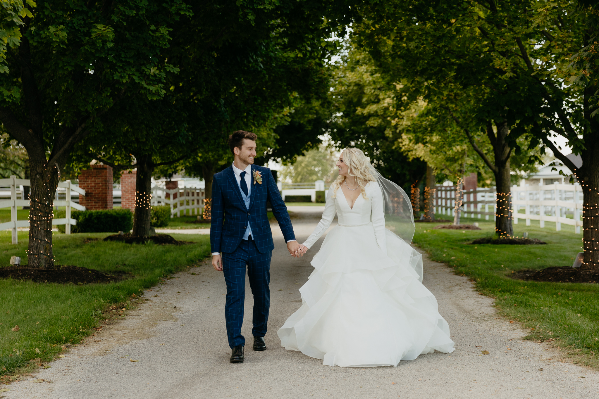 Bride and groom walk down tree lined driveway hand in hand