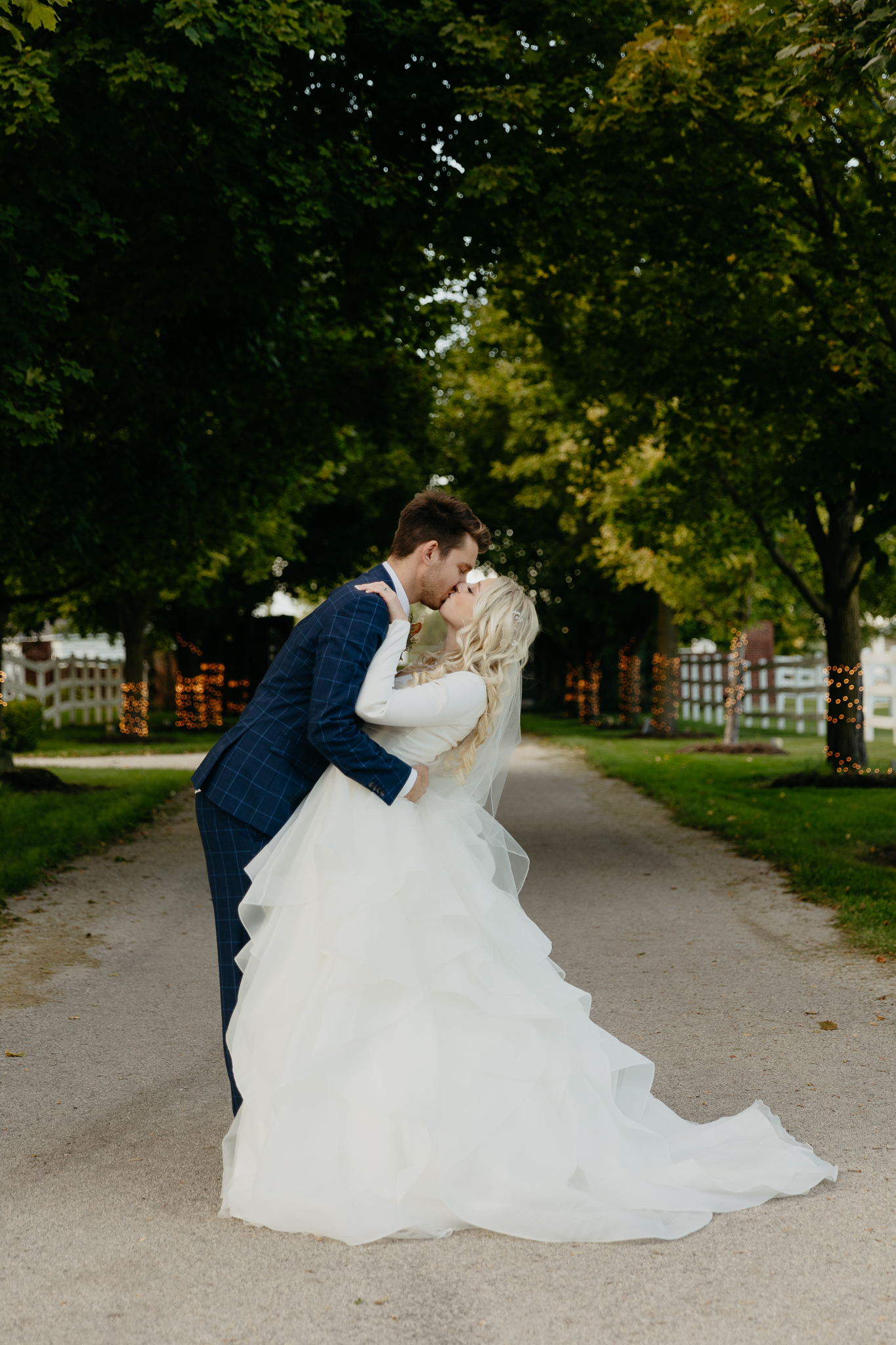 Bride and groom walk down tree lined driveway hand in hand while looking back, then kiss