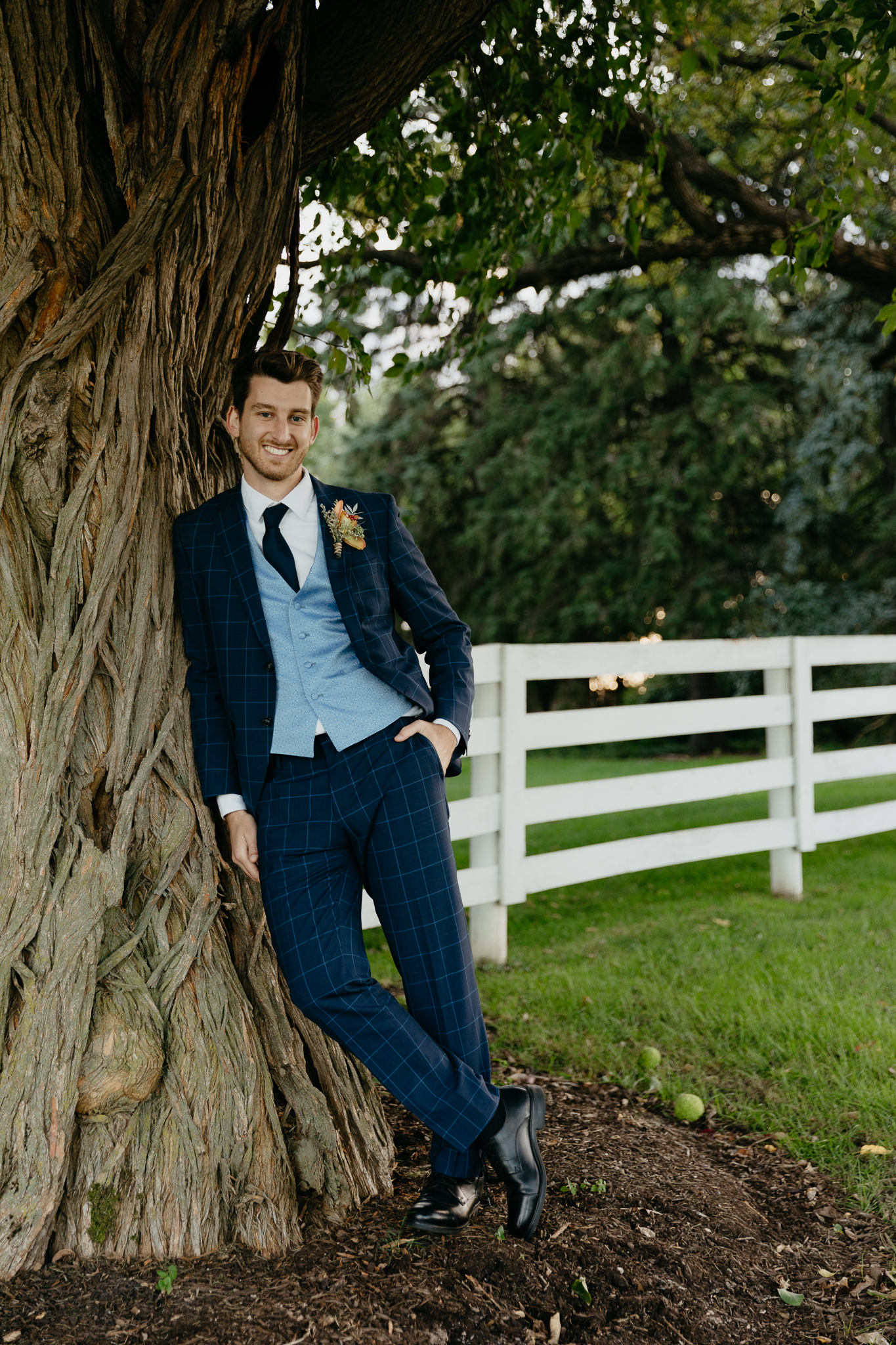 Groom with navy suit leans against a large tree with one hand in his pocket, smiling