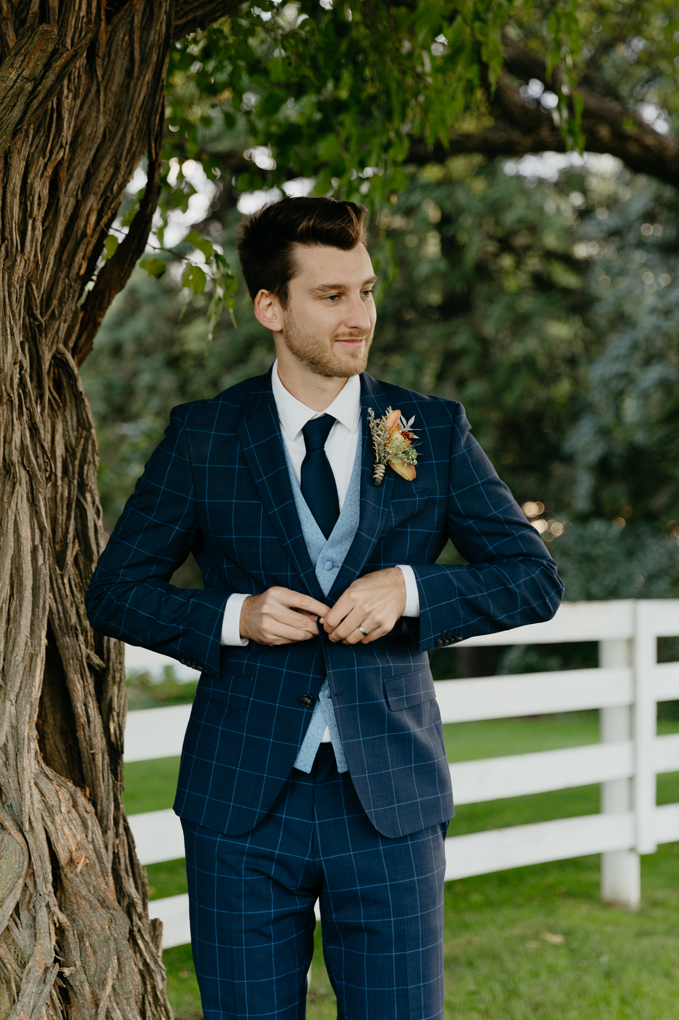 Groom adjusts his navy blue suit next to a horse pasture and large old tree
