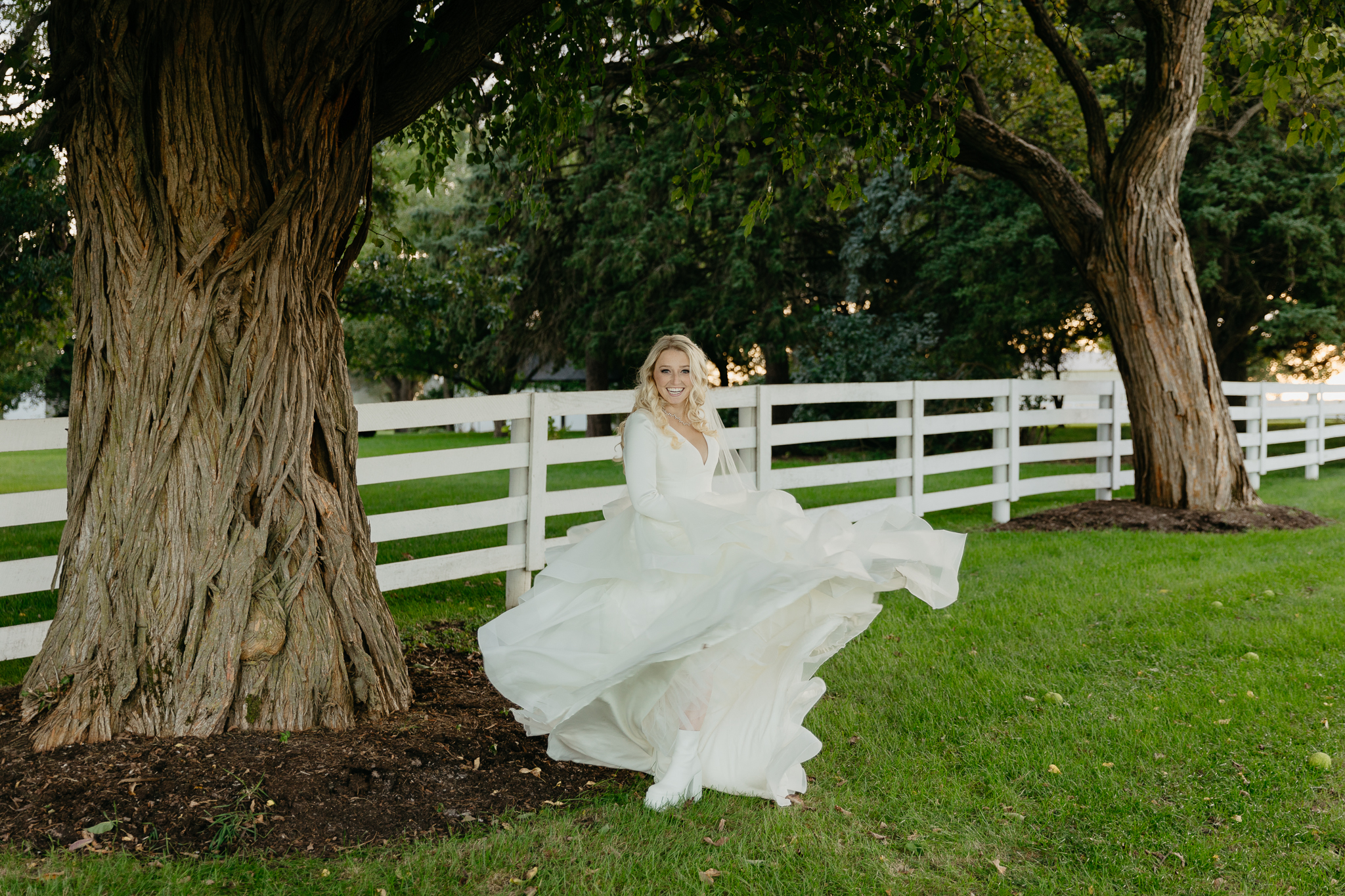 Bride twirls dress with white boots and laughs, underneath a large tree and next to a horse pasture
