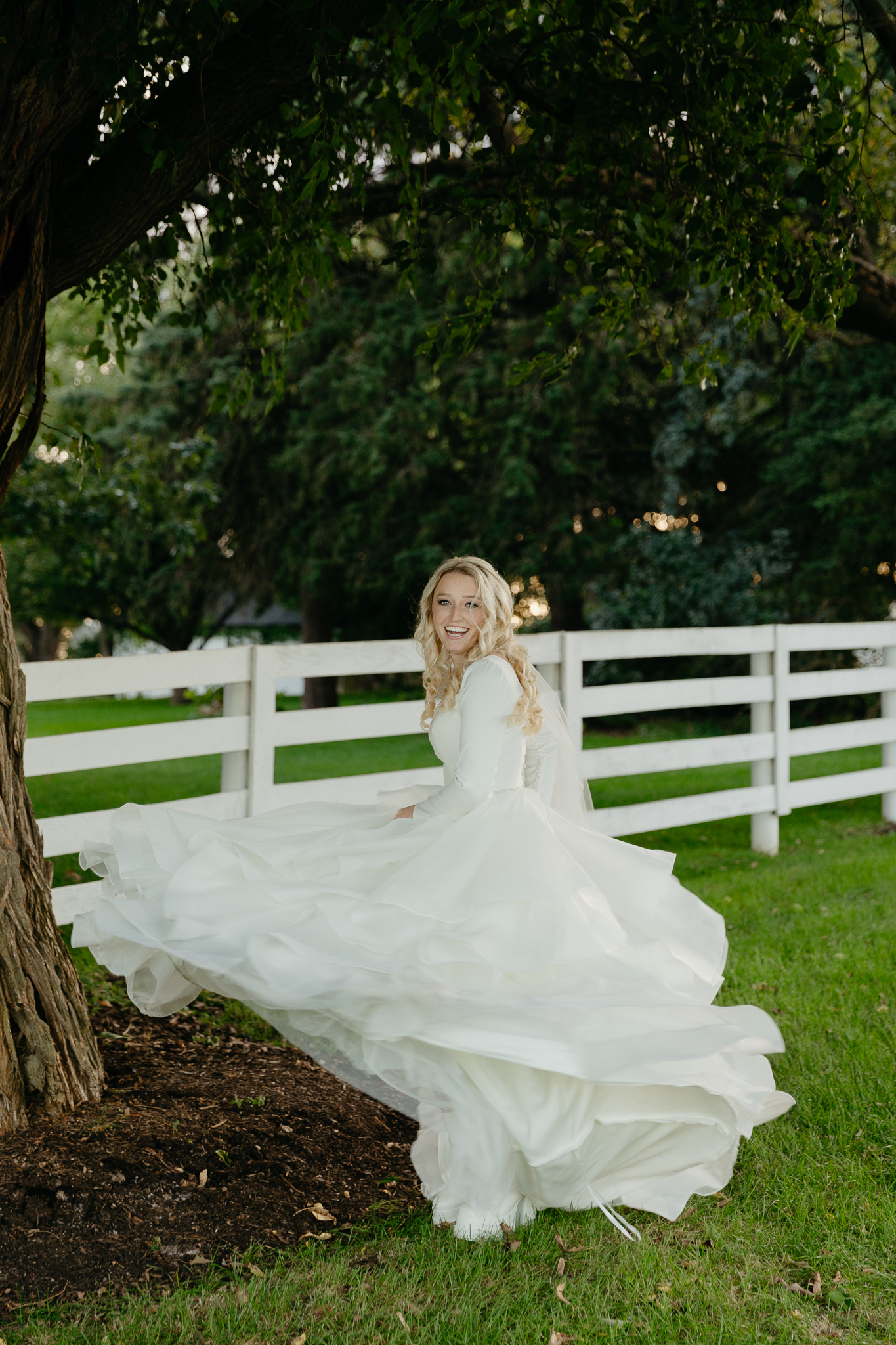Bride twirls dress with white boots and laughs, underneath a large tree and next to a horse pasture