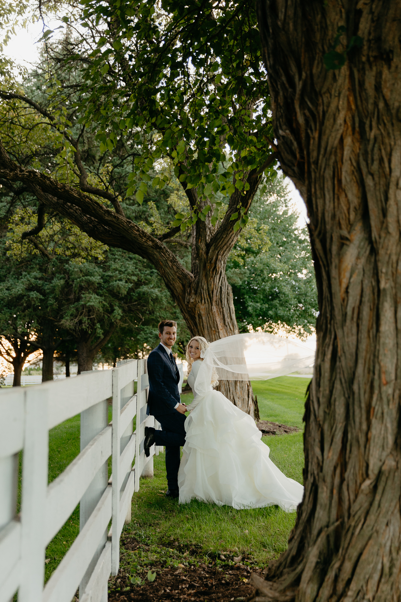 Bride and groom hold hands and lean against a white horse fence, smiling at the camera