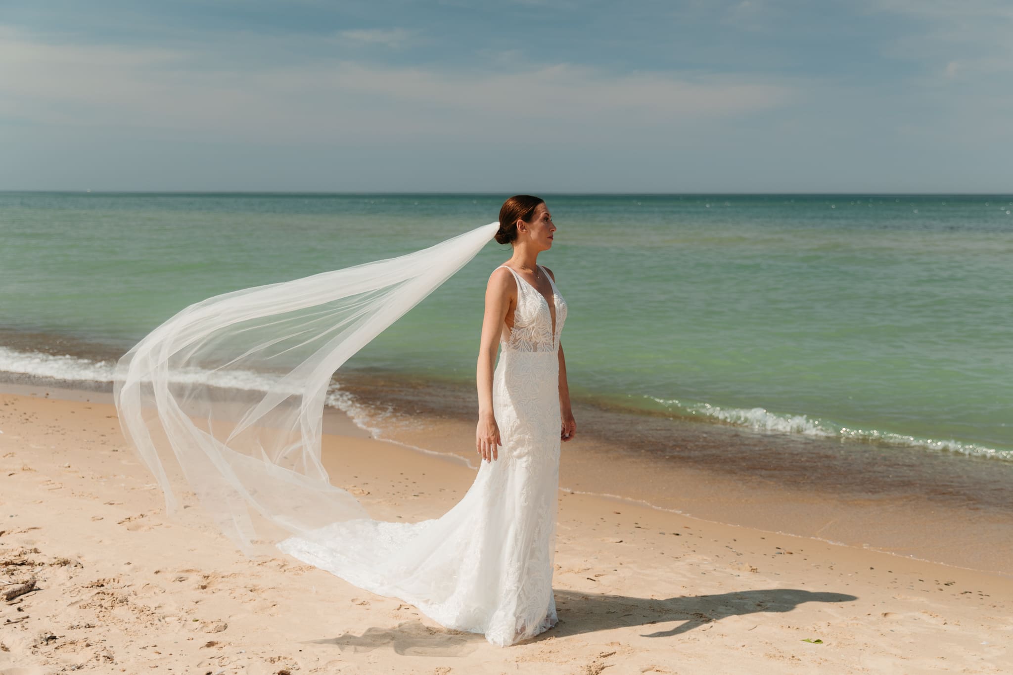 Bride poses next to the shoreline of Lake Michigan with her wedding veil blowing in the wind 