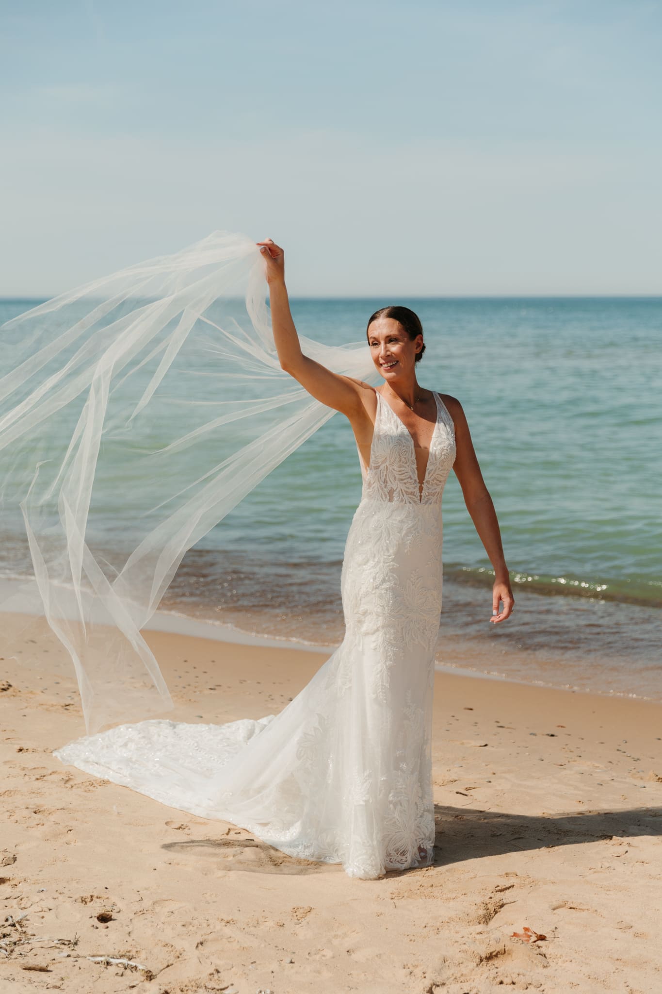 Bride poses next to the shoreline of Lake Michigan with her wedding veil blowing in the wind 