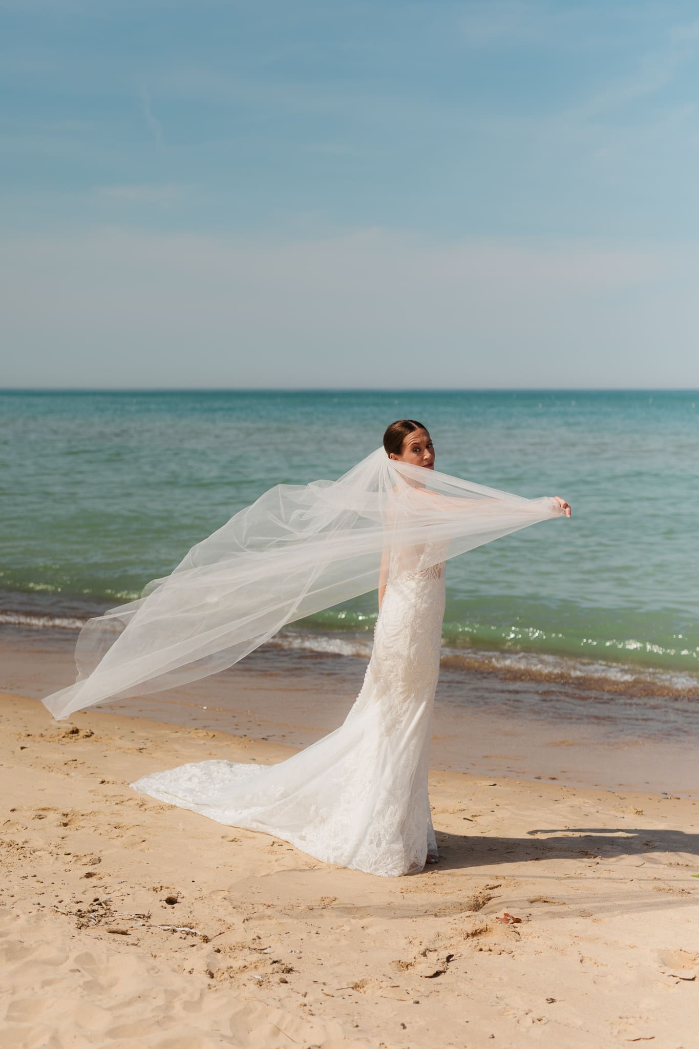 Bride poses next to the shoreline of Lake Michigan with her wedding veil blowing in the wind 
