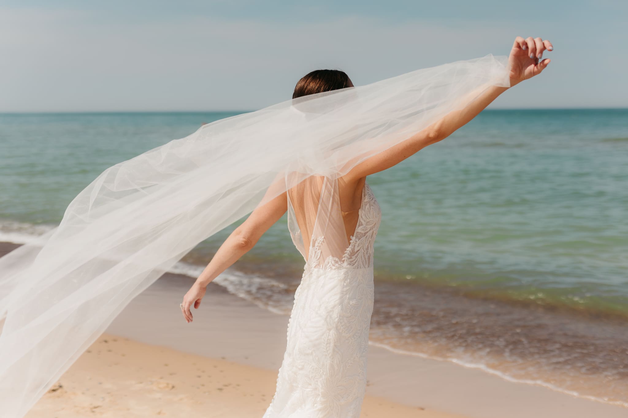 Bride poses next to the shoreline of Lake Michigan with her wedding veil blowing in the wind 