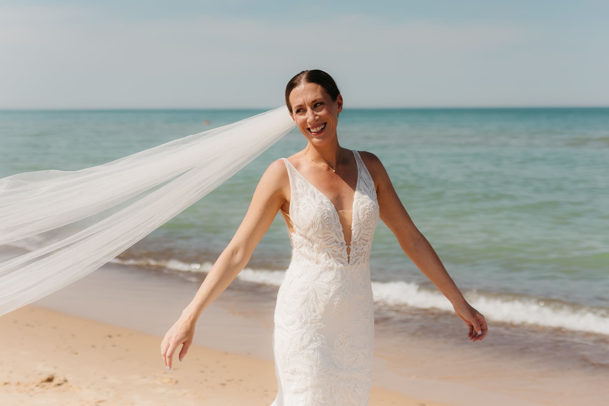 Bride poses next to the shoreline of Lake Michigan with her wedding veil blowing in the wind 