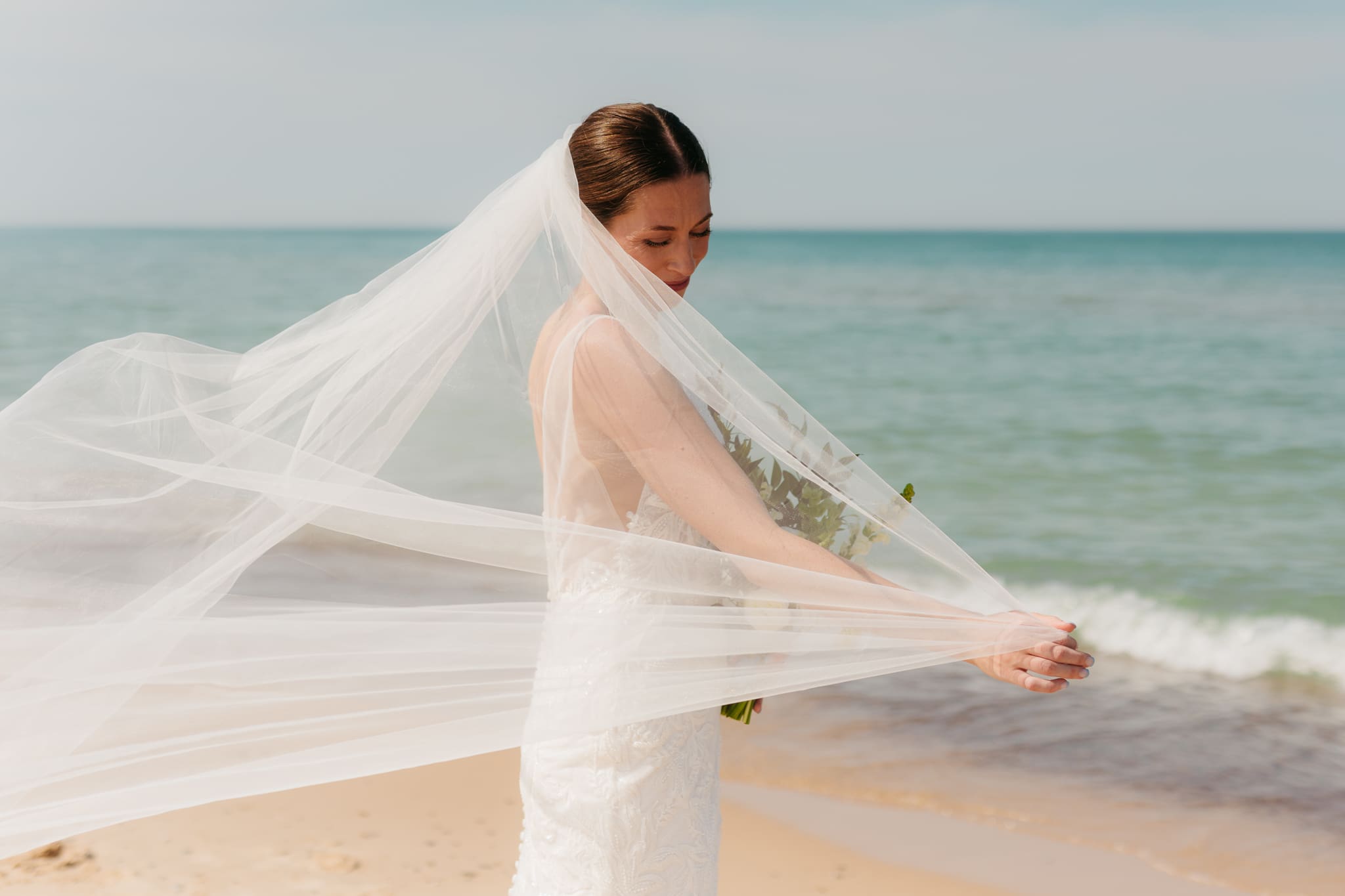 Bride poses next to the shoreline of Lake Michigan with her wedding veil blowing in the wind 