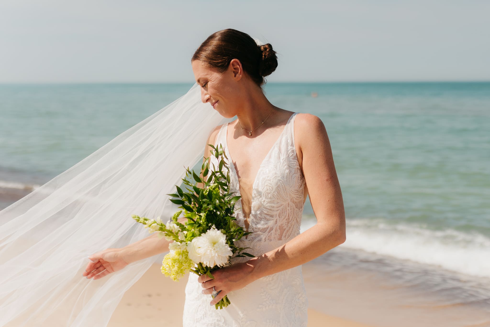 Bride poses next to the shoreline of Lake Michigan with her wedding veil blowing in the wind 