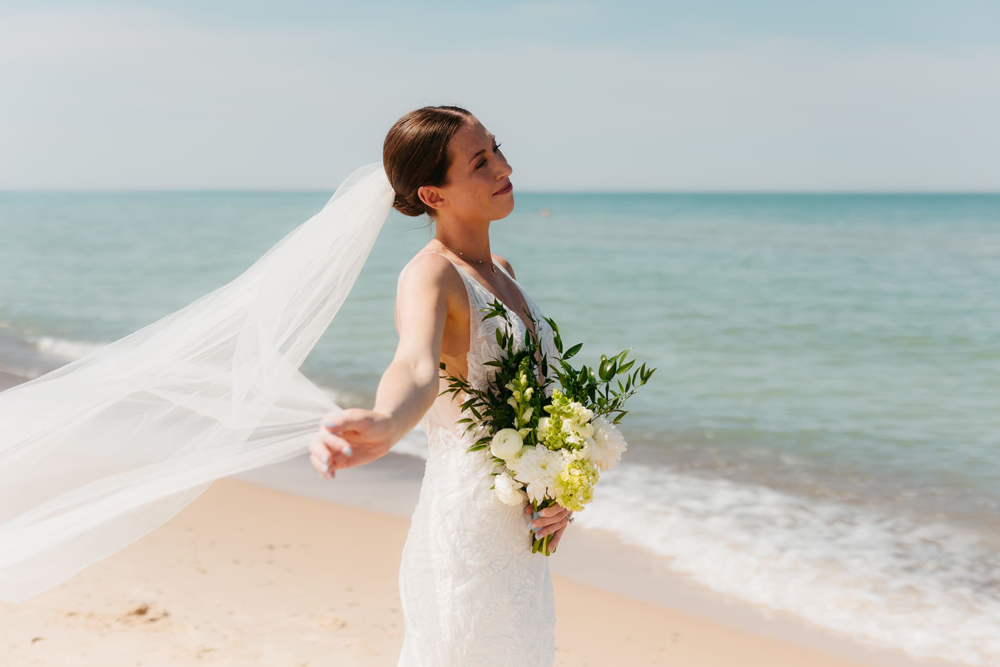 Bride poses next to the shoreline of Lake Michigan with her wedding veil blowing in the wind 