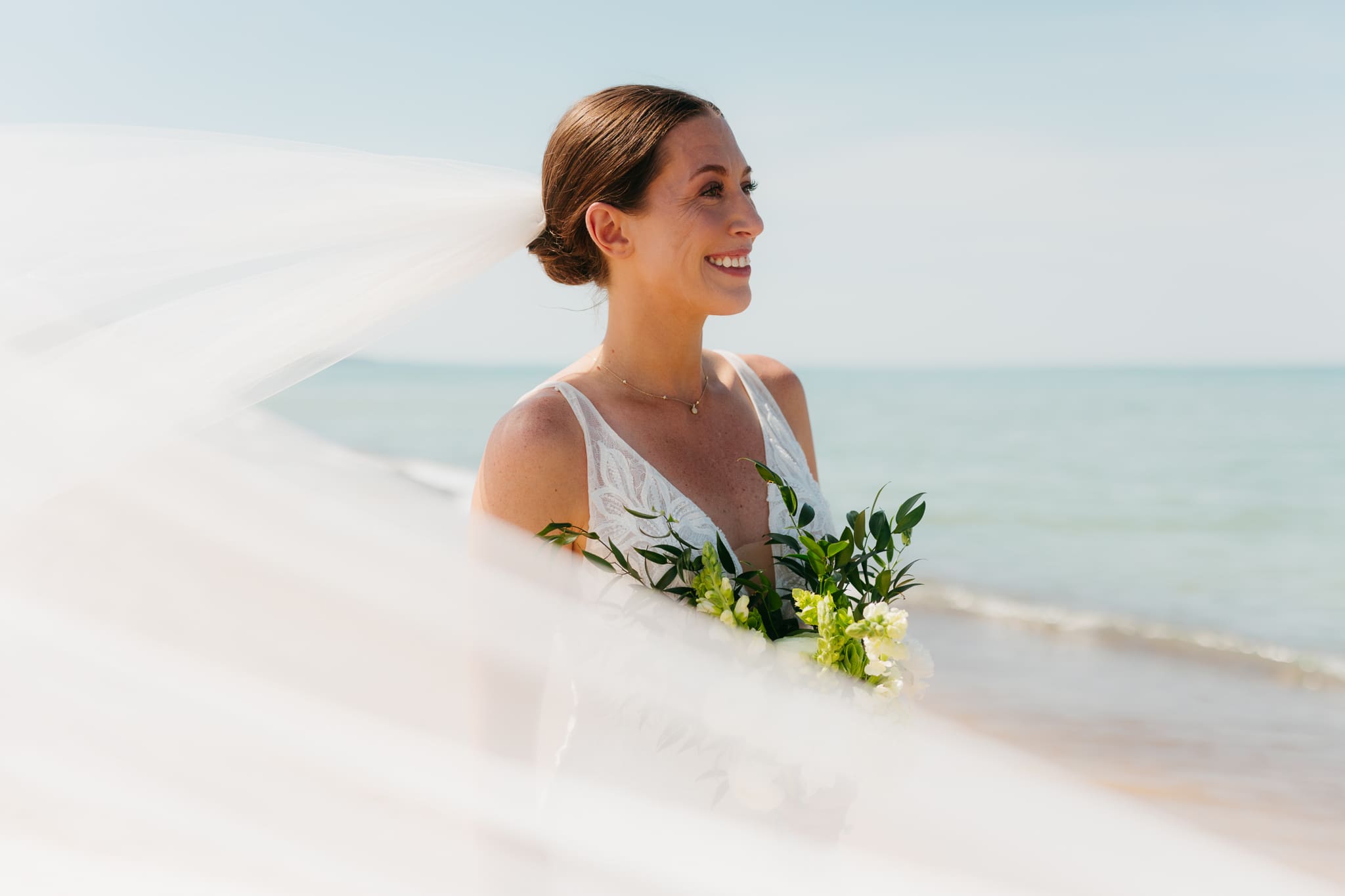 Bride poses next to the shoreline of Lake Michigan with her wedding veil blowing in the wind 