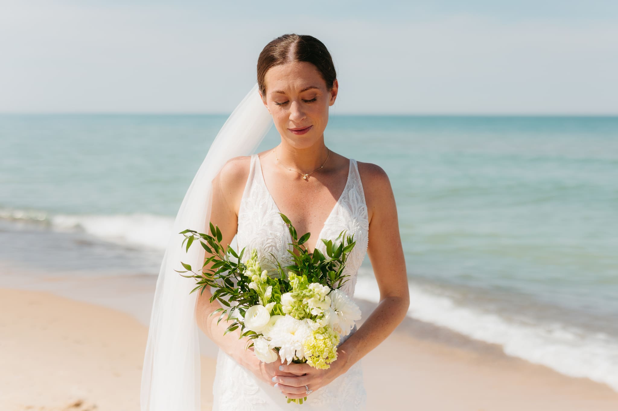 Bride poses next to the shoreline of Lake Michigan with her wedding veil blowing in the wind 