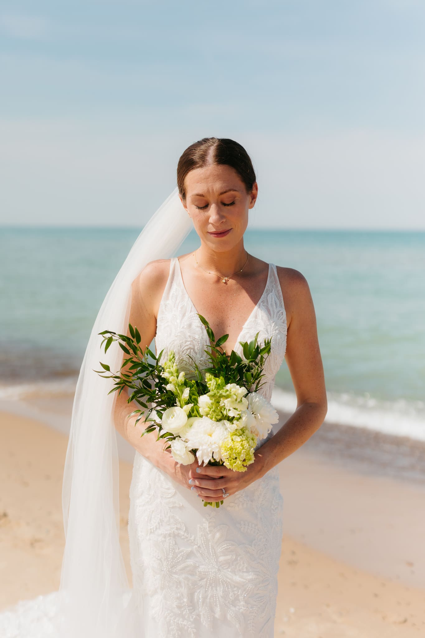 Bride poses next to the shoreline of Lake Michigan with her wedding veil blowing in the wind 