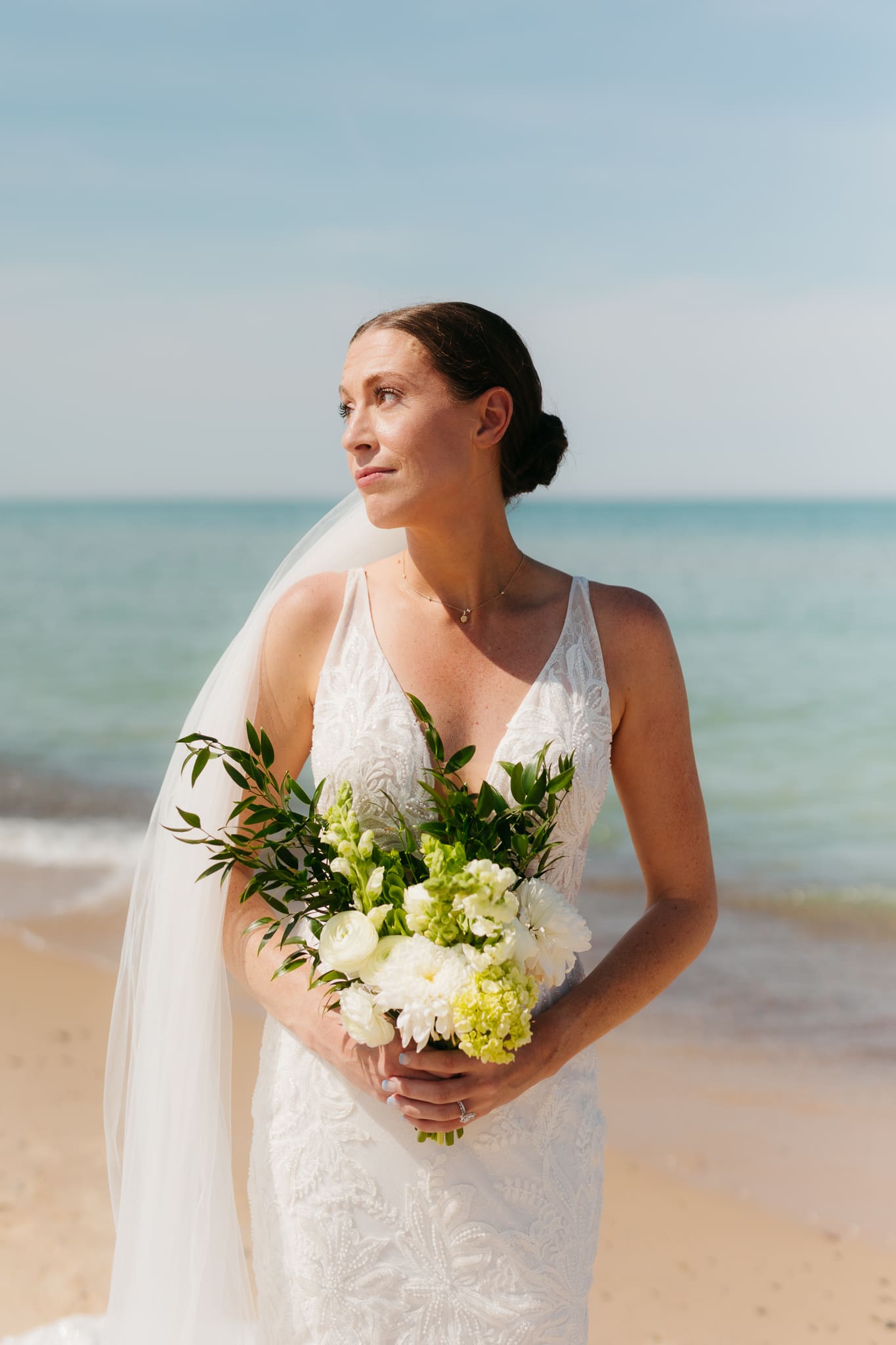 Bride poses next to the shoreline of Lake Michigan with her wedding veil blowing in the wind 