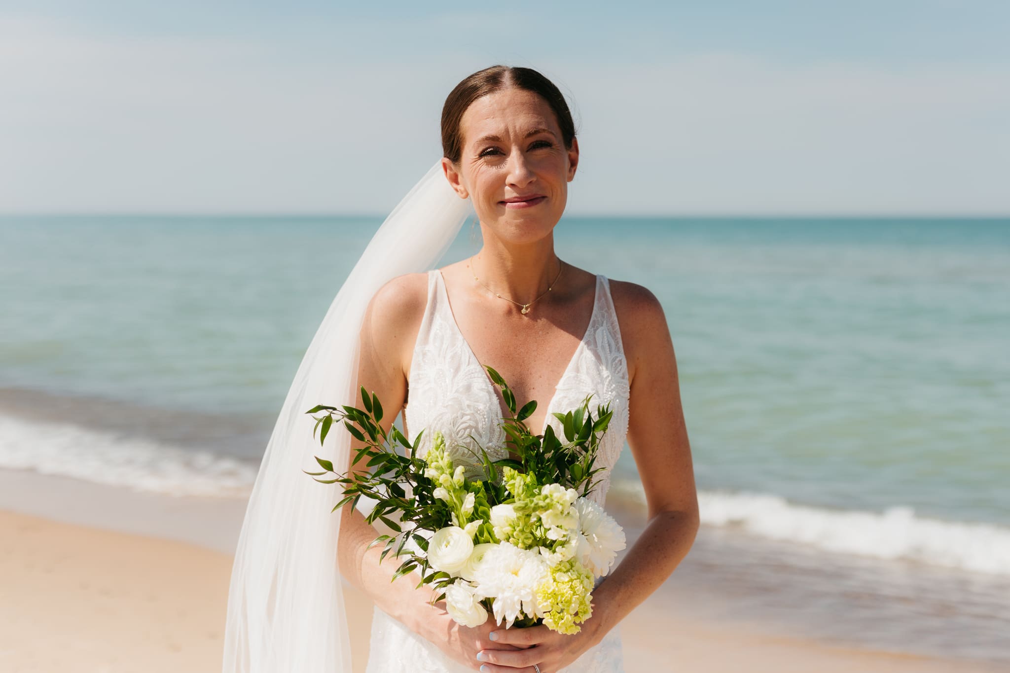 Bride poses next to the shoreline of Lake Michigan with her wedding veil blowing in the wind 