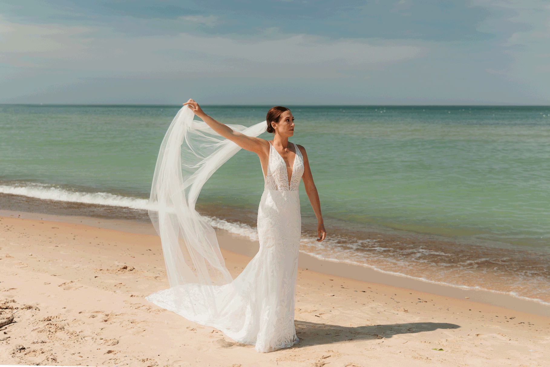 Bride poses next to the shoreline of Lake Michigan with her wedding veil blowing in the wind 